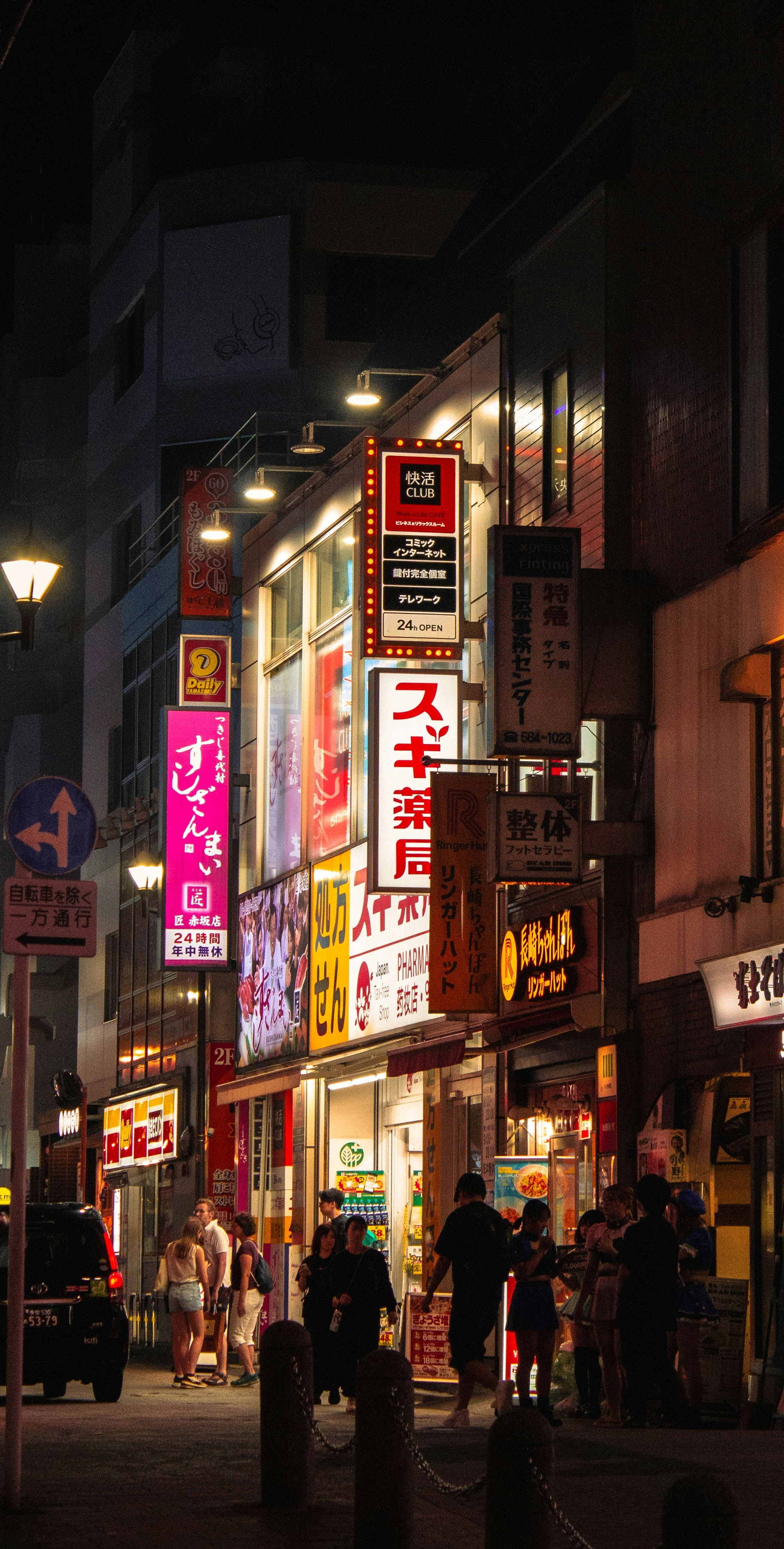 A Tokyo backstreet after midnight stacked with glowing signage, a few figures drifting past softly lit storefronts.
